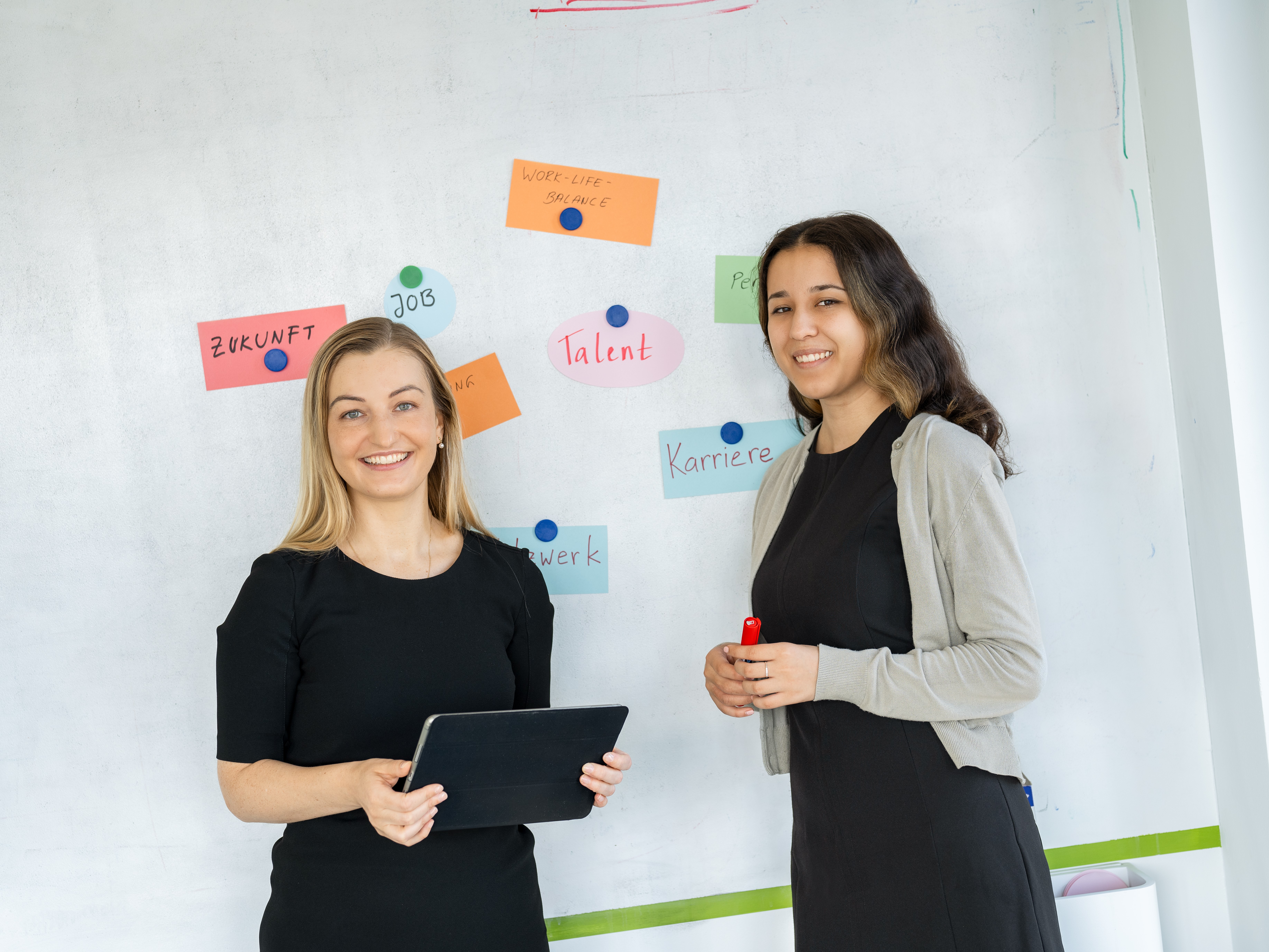 Two women are standing in front of a white board.