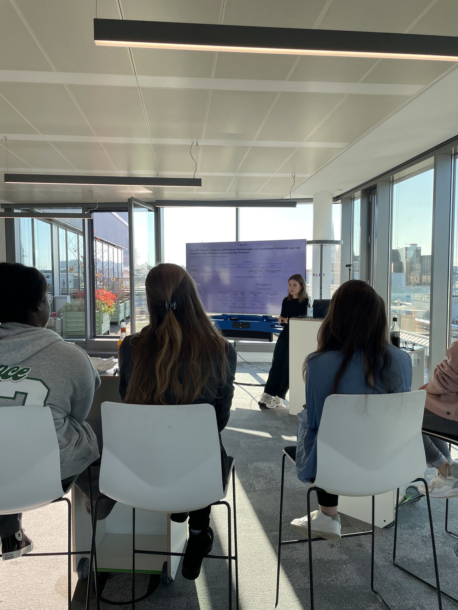 a group of people sitting in chairs in a room with a woman standing in front of a screen