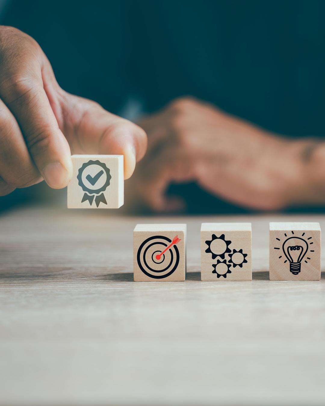 Hand places a wooden block with a quality symbol next to other blocks featuring a target, gears, and a light bulb.