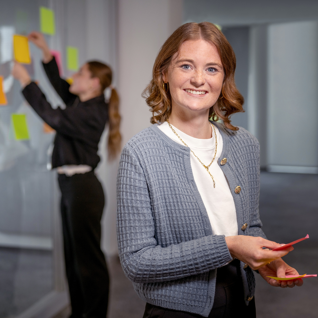 A young woman in the foreground is holding two Post-it stamps, and in the background another woman is sticking them onto a glass wall. 