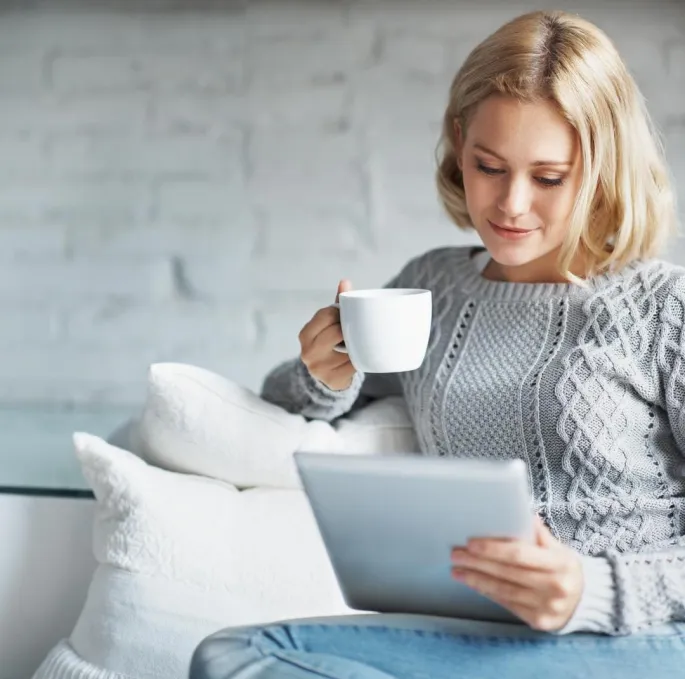 Eine Frau sitzt auf einem Sofa und hält eine Kaffeetasse und ein Tablet in den Händen.