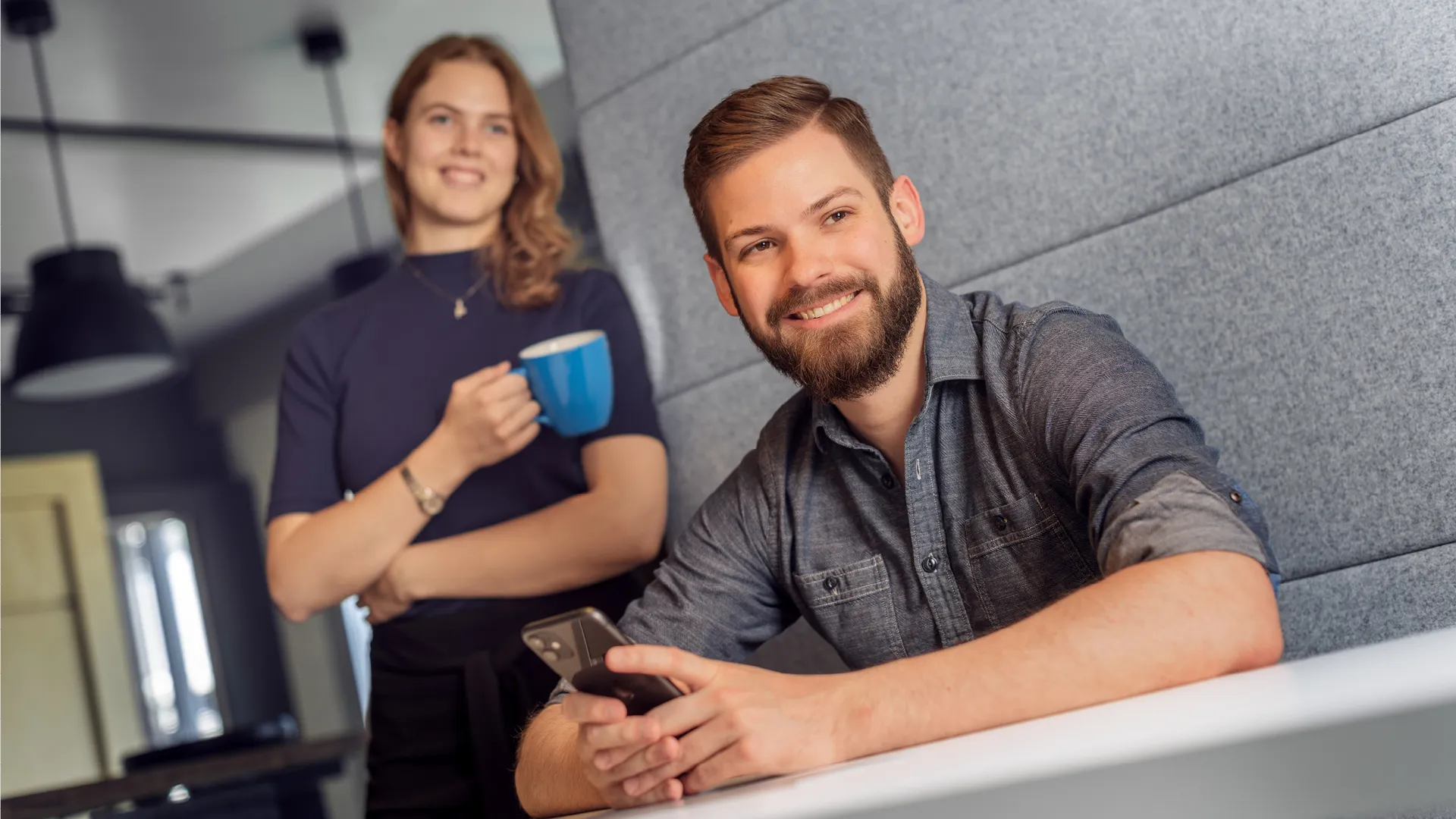 Zwei zeb-Mitarbeitende sind zu sehen. Einer sitzt am Tisch mit seinem Handy, die andere steht mit einer zeb-Tasse im Hintergrund.