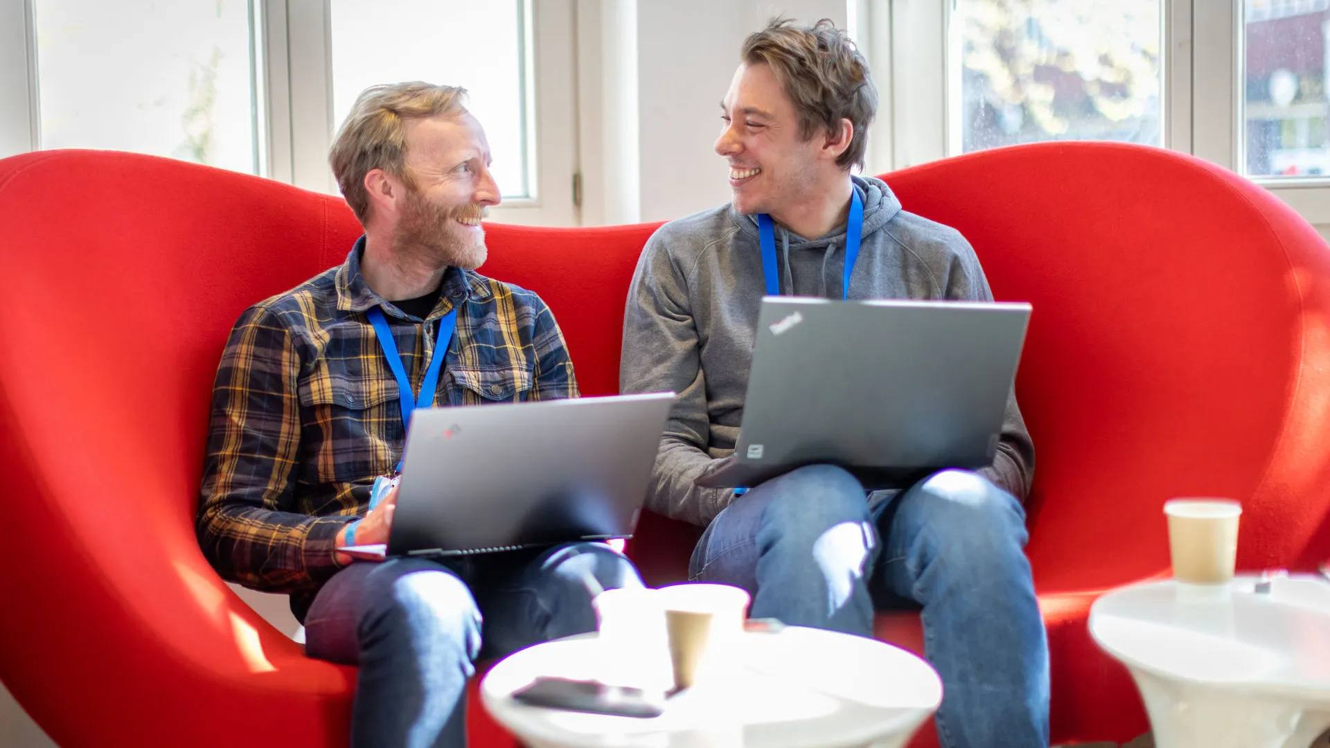 Zwei Kollegen mit Laptop lächelnd auf rotem Sofa