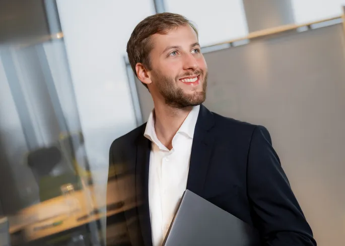 Ein Mitarbeiter von zeb hält einen Laptop unter dem Arm. Im Hintergrund befindet sich eine Tafel.
