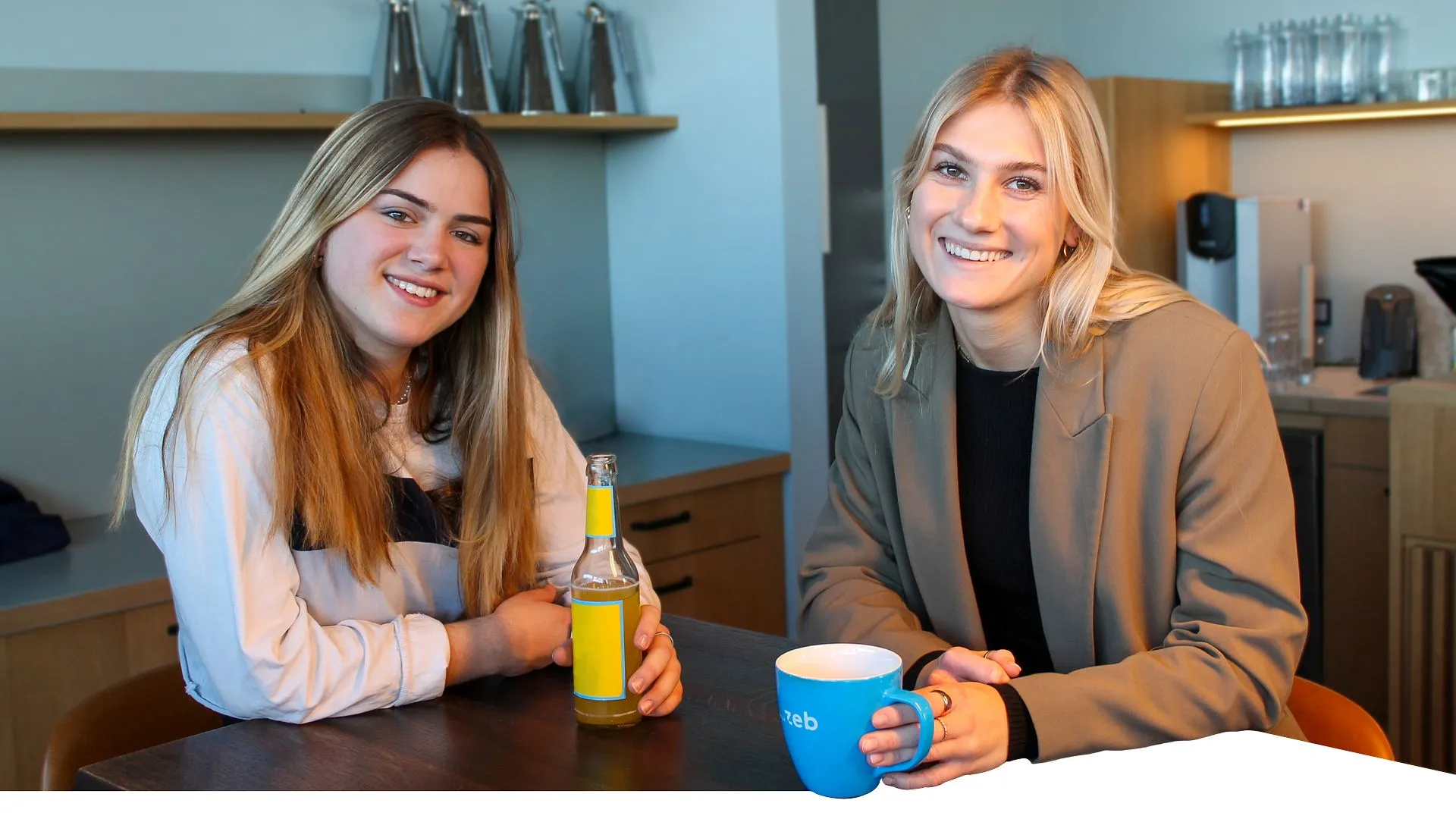Two young women are drinking coffee and are smiling into the camera