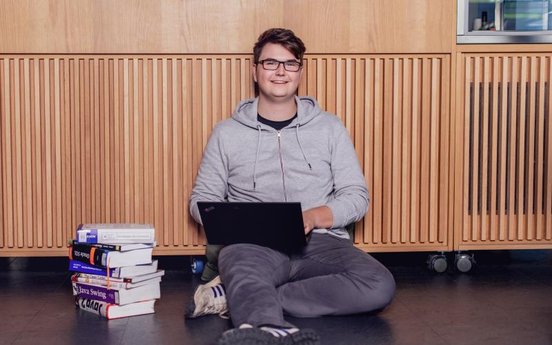 Ein Student lehnt an einer Wand und sitzt mit seinem Laptop auf dem Schoß auf dem Boden. Neben ihm steht ein Stapel Bücher.