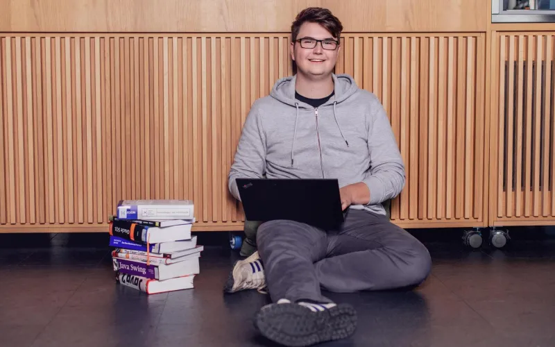 Ein Mann sitzt neben einem Stapel Bücher und mit seinem Laptop auf dem Boden. 