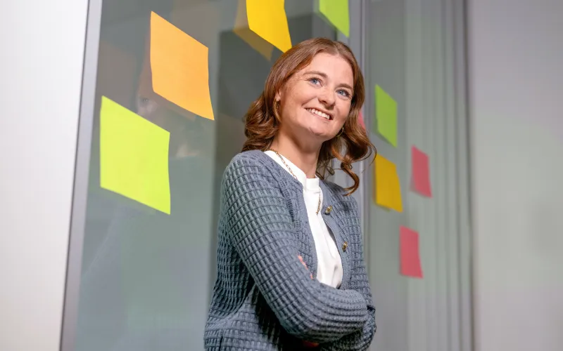 Eine Frau lächelt mit verschränkten Armen und gelben Post-its an einer Glaswand