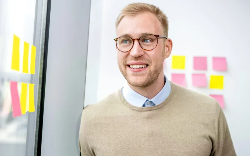 Ein Mann trägt eine Brille und einen beigen Pullover. Er schaut aus dem Fenster.
