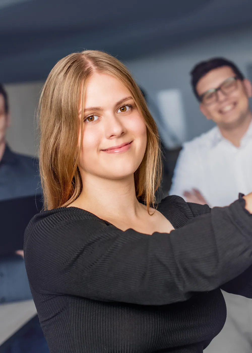 Eine Studentin klebt ein Post-It an eine Tafel. Im Hintergrund stehen zwei weitere Studenten und lächeln in die Kamera.